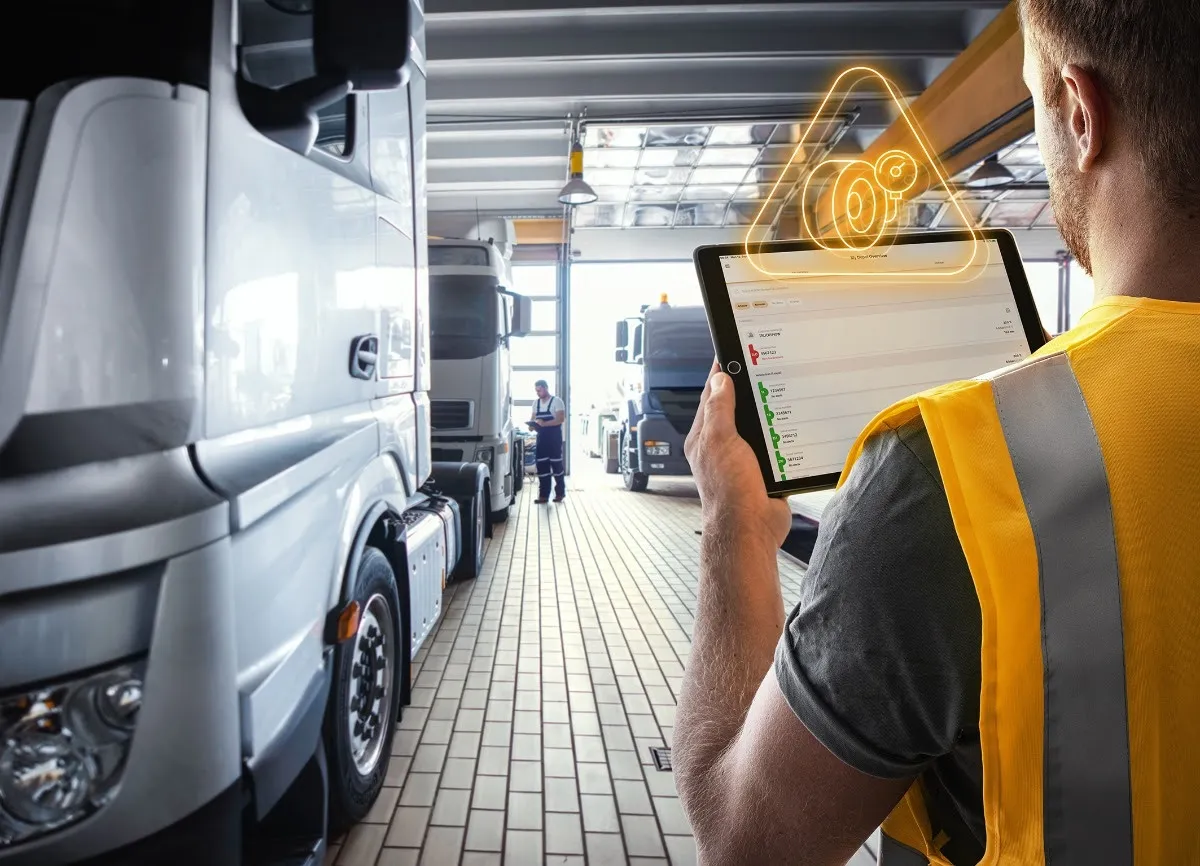 Image of the back of a man in a high-vis vest holding a tablet while near a white lorry on the left