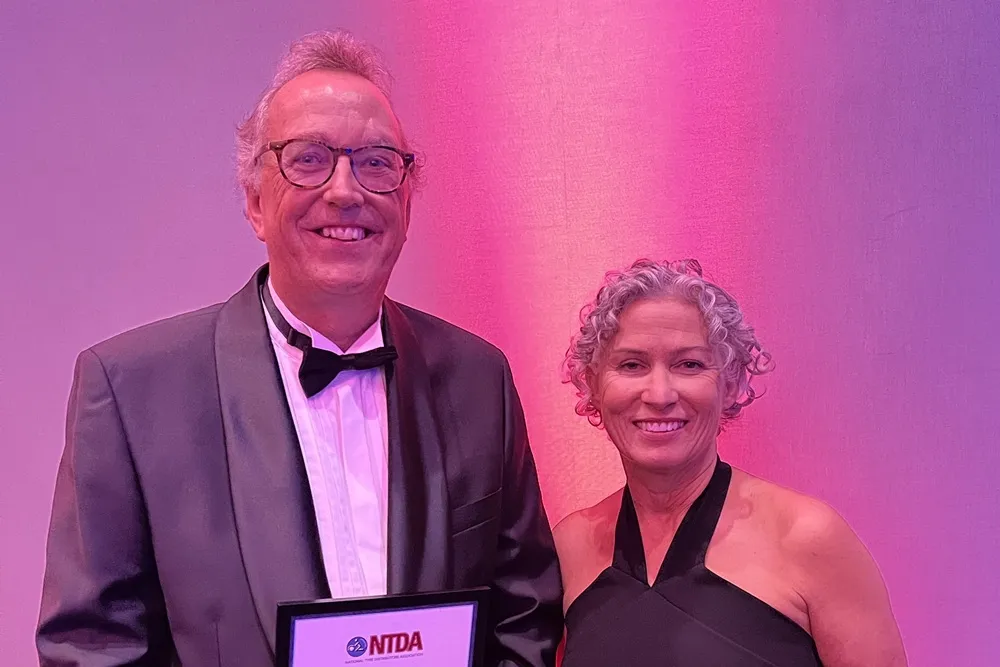 Image of Steve Howet on the left, and Tracey Mortimer on the right at the NTDA Awards with a pink/purple background